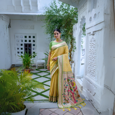 Woman wearing a yellow silk saree with green blouse and gold border, featuring a handpainted pallu with peacock and floral design, posing in a white heritage courtyard with potted plants and traditional architectural details.