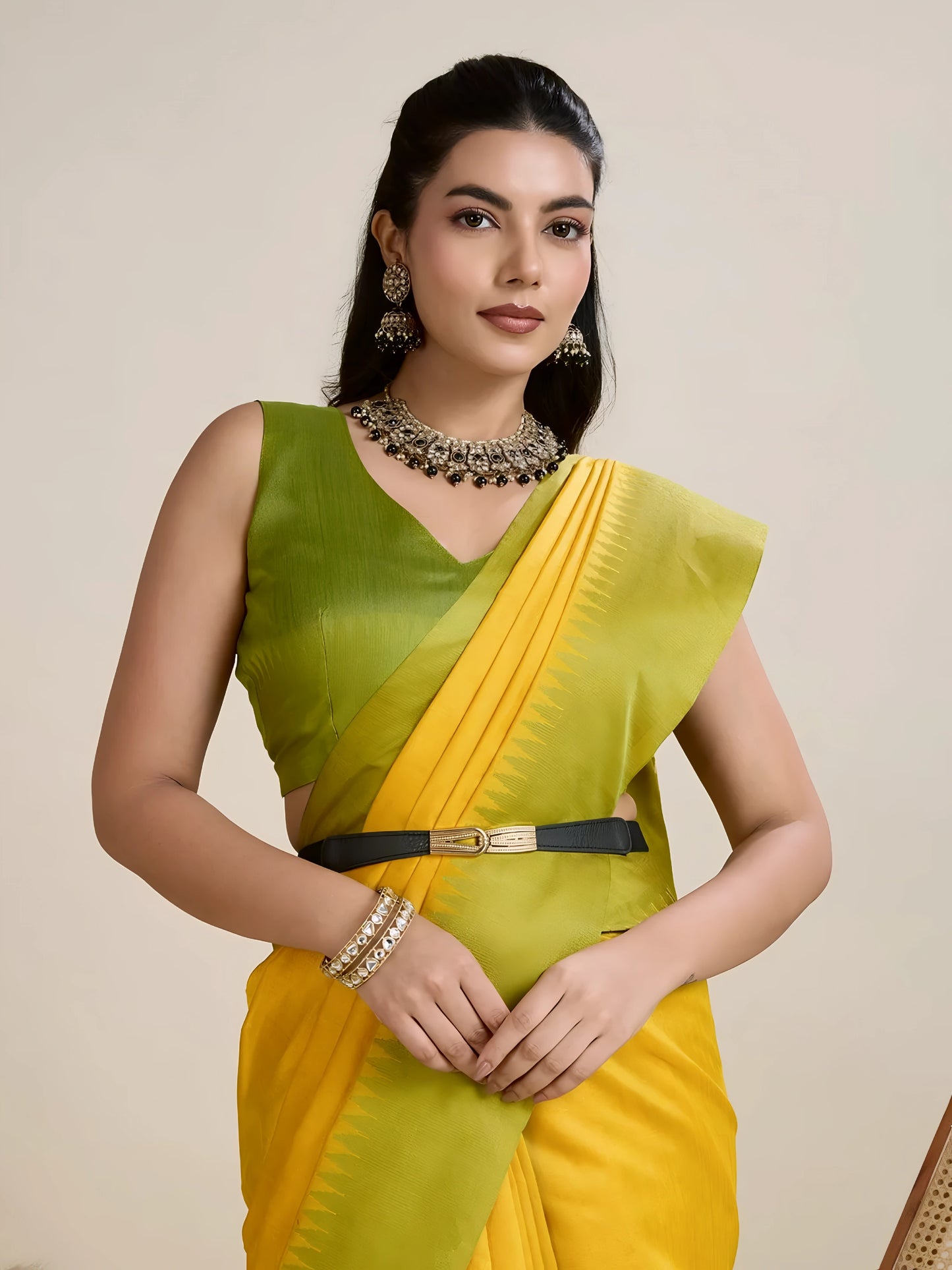 Woman wearing a yellow silk saree with a contrasting green border and pallu, paired with a matching blouse and traditional jewelry, elegantly posing beside a wooden chair in a minimalist studio setting with neutral decor, ceramic vases, and pampas grass.