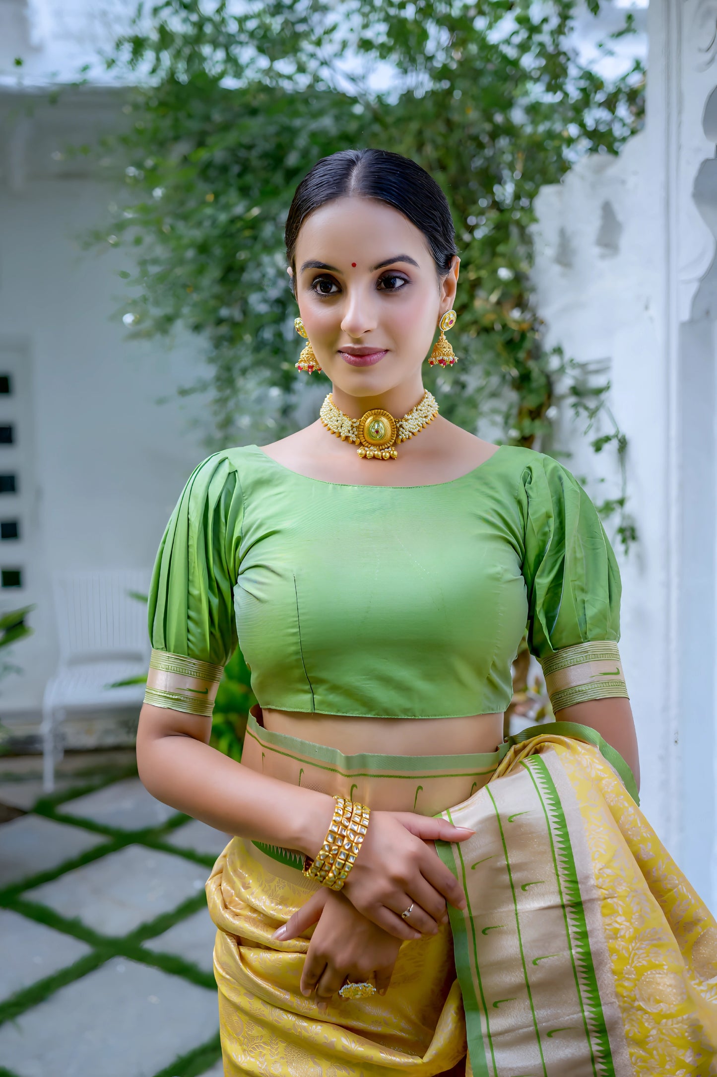 Woman wearing a yellow silk saree with green blouse and gold border, featuring a handpainted pallu with peacock and floral design, posing in a white heritage courtyard with potted plants and traditional architectural details