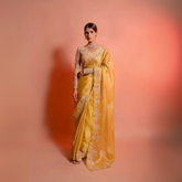Woman wearing a yellow silk saree with floral embroidery and a matching embellished blouse, posing in a studio with a peach gradient background.