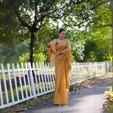 Woman wearing a mustard yellow silk saree with subtle floral embroidery and a contrasting maroon blouse, accessorized with traditional jewelry and bangles, standing beside a white picket fence in a sunlit garden with lush greenery and trees in the background.
