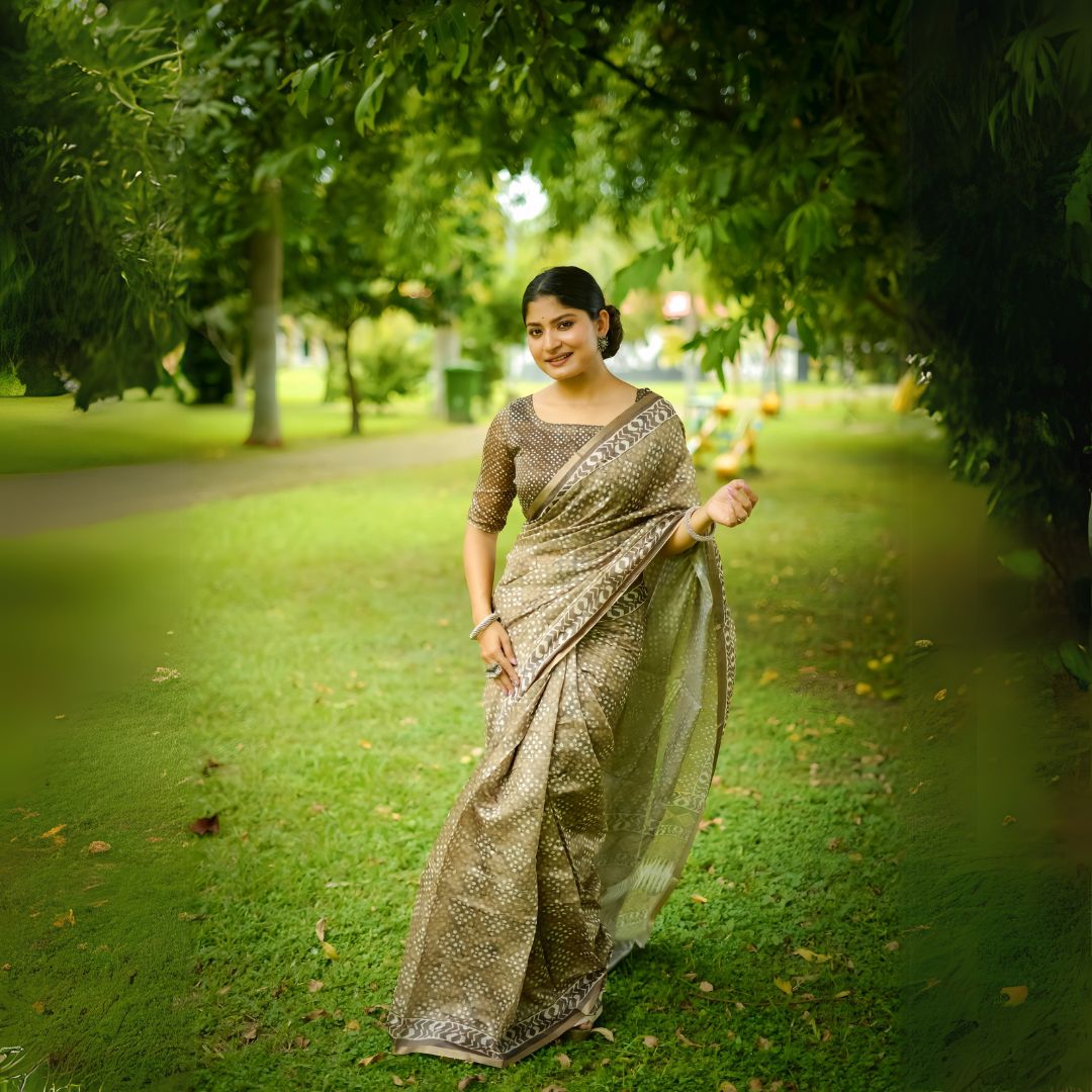 Woman wearing an Beige handloom saree with intricate geometric and dotted patterns, paired with a matching blouse, standing gracefully on green grass in a lush outdoor garden with trees in the background.