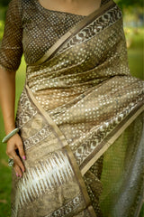 Woman wearing an Beige handloom saree with intricate geometric and dotted patterns, paired with a matching blouse, standing gracefully on green grass in a lush outdoor garden with trees in the background