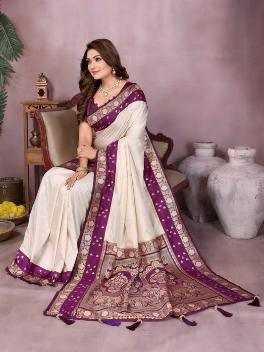 Woman wearing an ivory Banarasi saree with an ornate deep purple and gold embroidered border, featuring traditional woven motifs and a matching purple blouse with gold detailing. She poses gracefully in a modern studio with large white vases, a wooden table adorned with brass decor, green foliage, and a bowl of yellow flowers for an elegant setting.