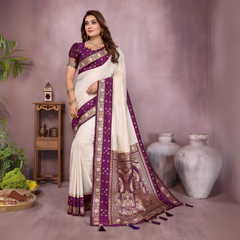 Woman wearing an ivory Banarasi saree with an ornate deep purple and gold embroidered border, featuring traditional woven motifs and a matching purple blouse with gold detailing. She poses gracefully in a modern studio with large white vases, a wooden table adorned with brass decor, green foliage, and a bowl of yellow flowers for an elegant setting.