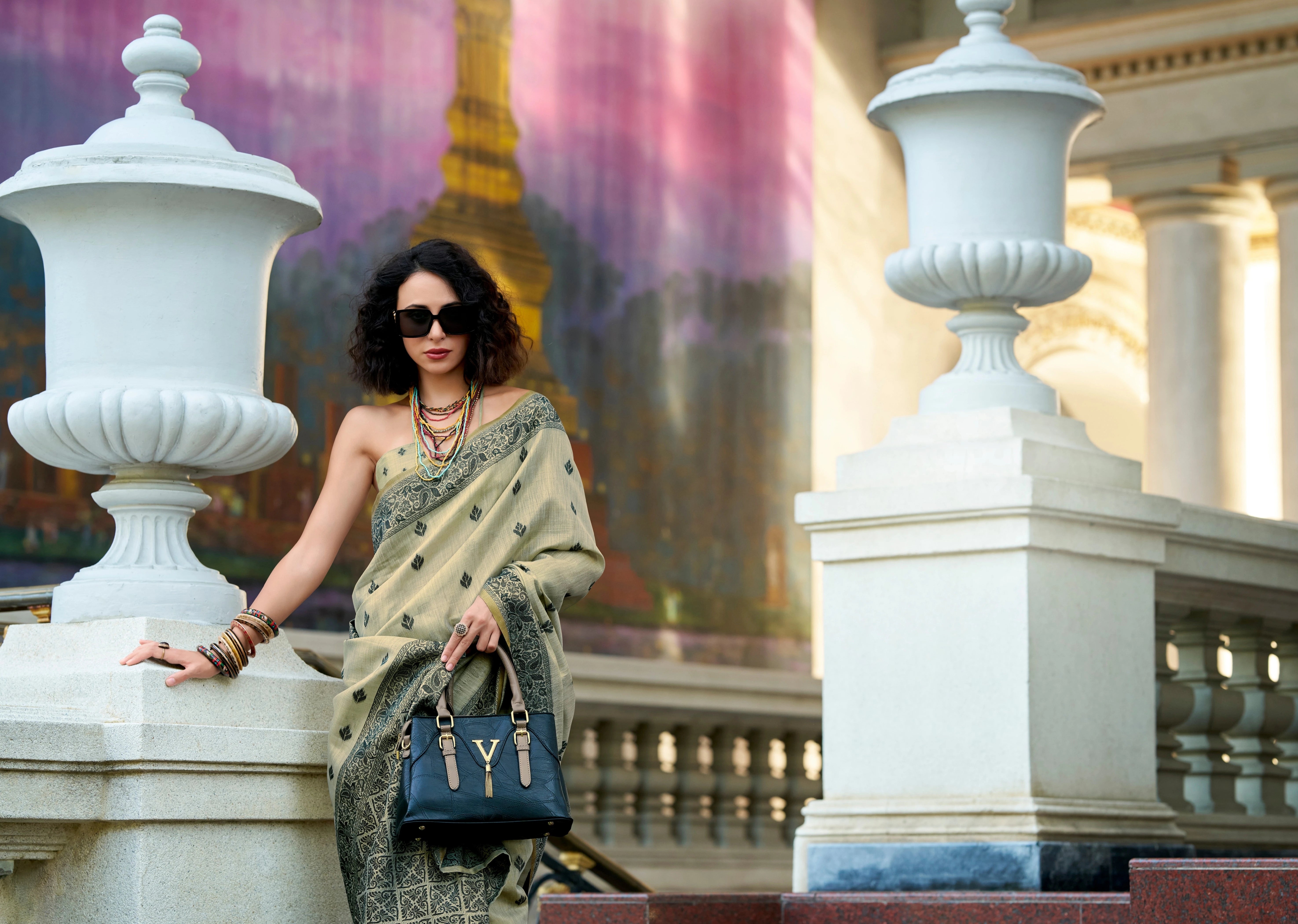 Woman in a saree with sunglasses and a handbag in front of a blurred background