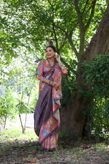 Woman wearing a mauve saree with vibrant Madhubani-inspired floral print border and pallu, paired with a matching blouse and traditional jewelry, standing gracefully on a garden pathway surrounded by lush green trees and foliage.
