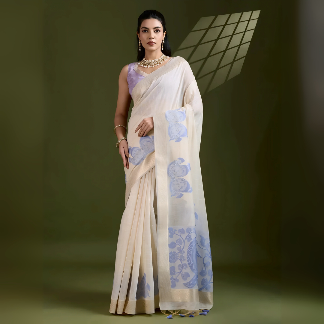 Woman wearing an ivory silk saree with blue floral motifs and a lavender blouse, accessorized with pearl jewelry, posing in a contemporary olive green studio with a geometric window shadow on the wall.
