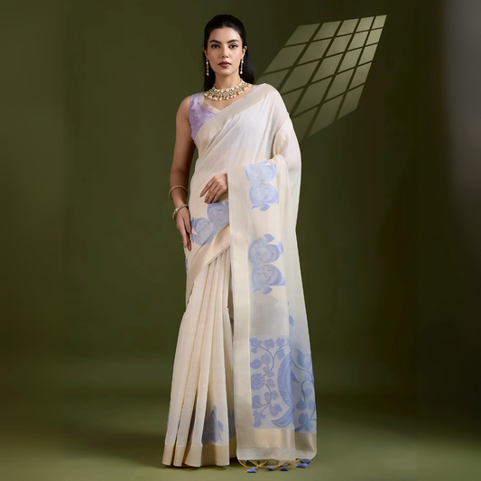 Woman wearing an ivory silk saree with blue floral motifs and a lavender blouse, accessorized with pearl jewelry, posing in a contemporary olive green studio with a geometric window shadow on the wall.
