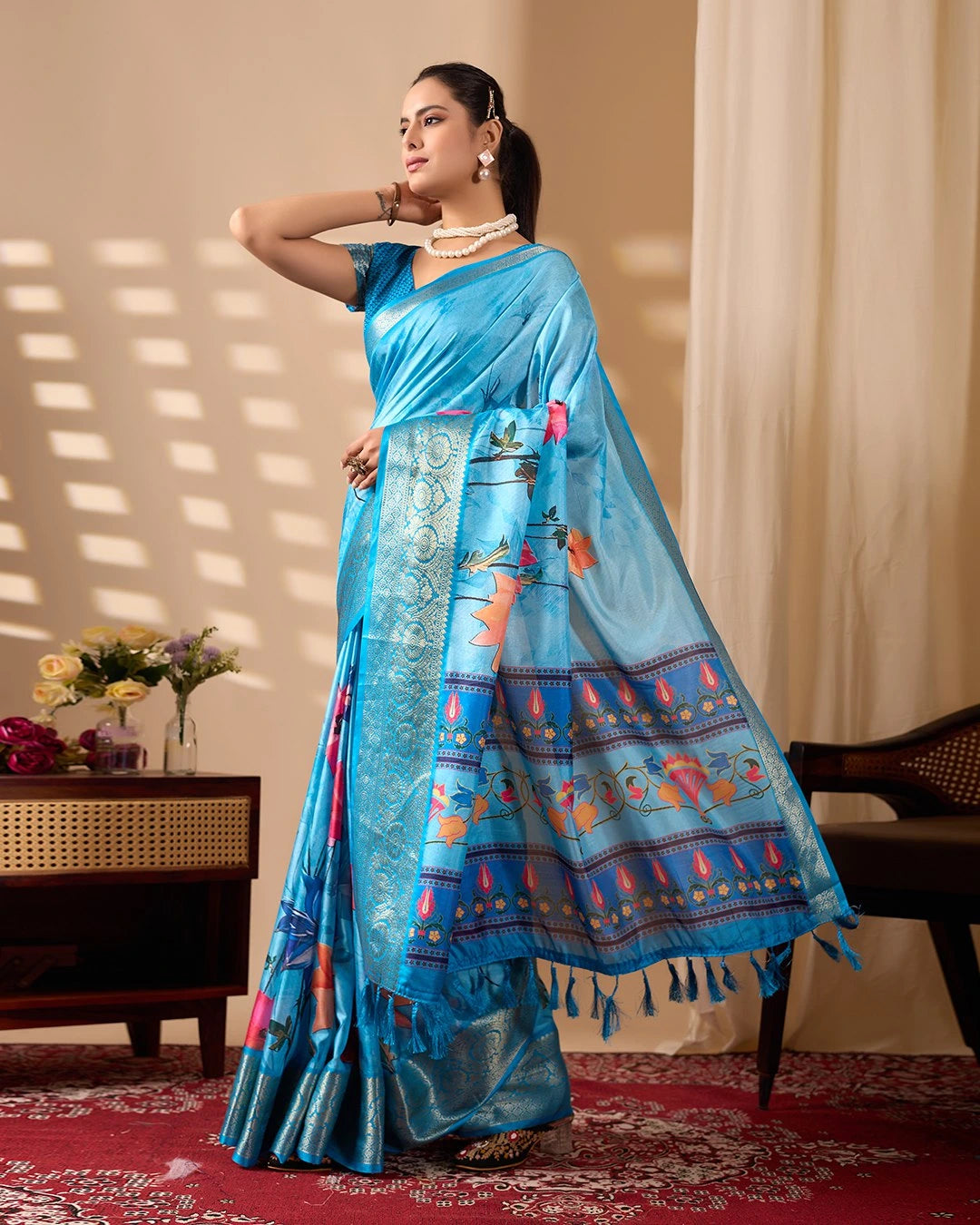 Woman wearing a blue saree with intricate patterns in a room with a table and flowers.