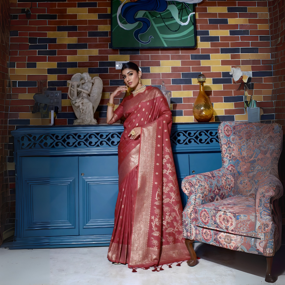 Woman wearing a maroon silk saree with intricate gold woven patterns and a decorative gold border, accessorized with traditional gold jewelry, elegantly posing in a vintage-inspired room featuring a blue cabinet, multicolored brick wall, artistic sculptures, and an ornate patterned armchair.