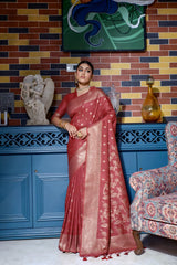 Woman wearing a maroon silk saree with intricate gold woven patterns and a decorative gold border, accessorized with traditional gold jewelry, elegantly posing in a vintage-inspired room featuring a blue cabinet, multicolored brick wall, artistic sculptures, and an ornate patterned armchair.