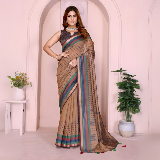 Woman wearing a beige silk saree adorned with subtle multicolor stripes and small dot patterns, paired with a sleeveless dark blouse and traditional jewelry including a statement necklace, bangles, and rings. She stands gracefully in a contemporary studio setting featuring a pastel arched alcove, a white side table with a floral vase, and a large potted green plant, showcasing elegant and understated ethnic style.