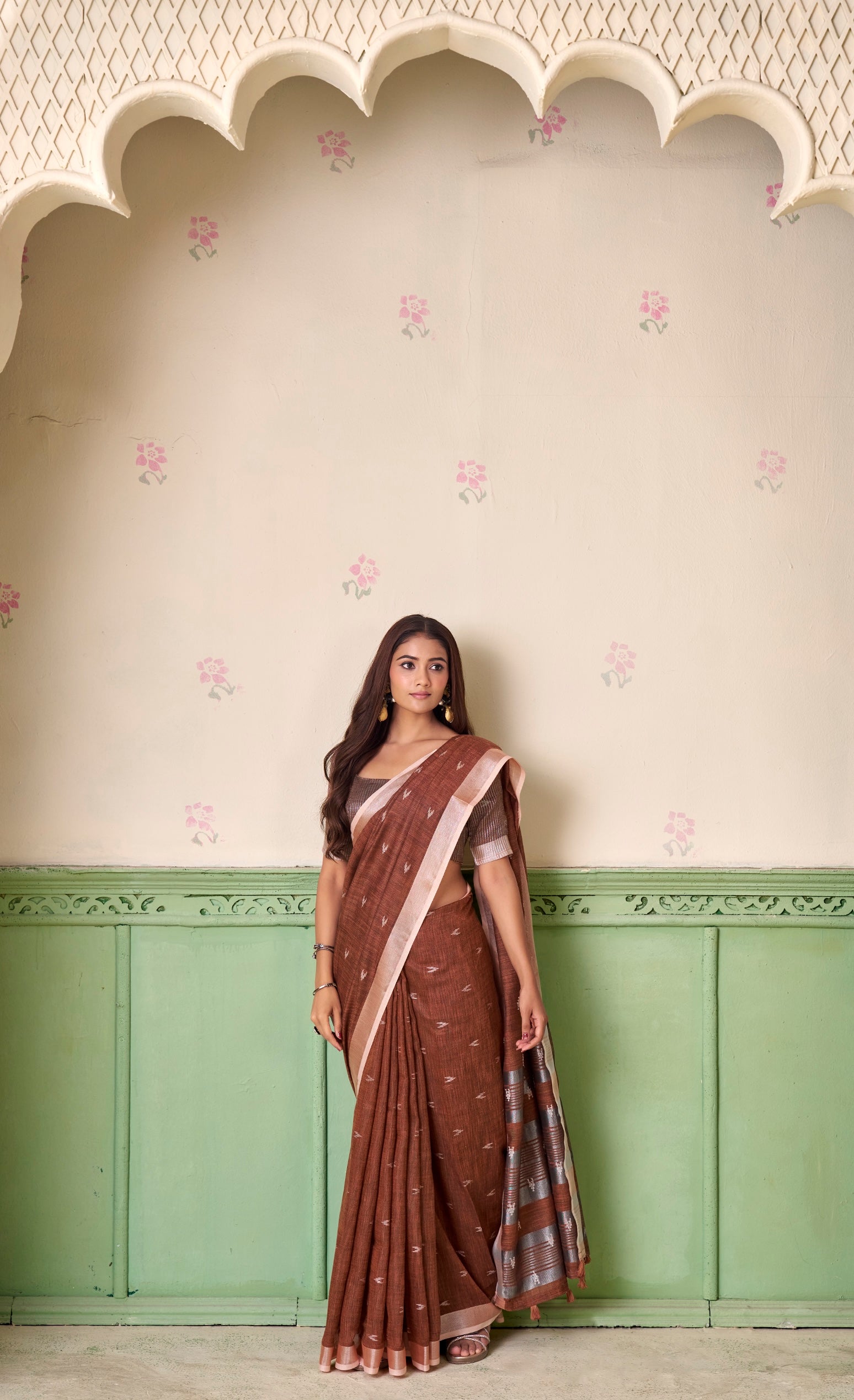 Woman wearing a brown saree with a pink border against a decorative wall.