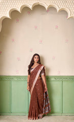 Woman wearing a brown saree with a pink border against a decorative wall.