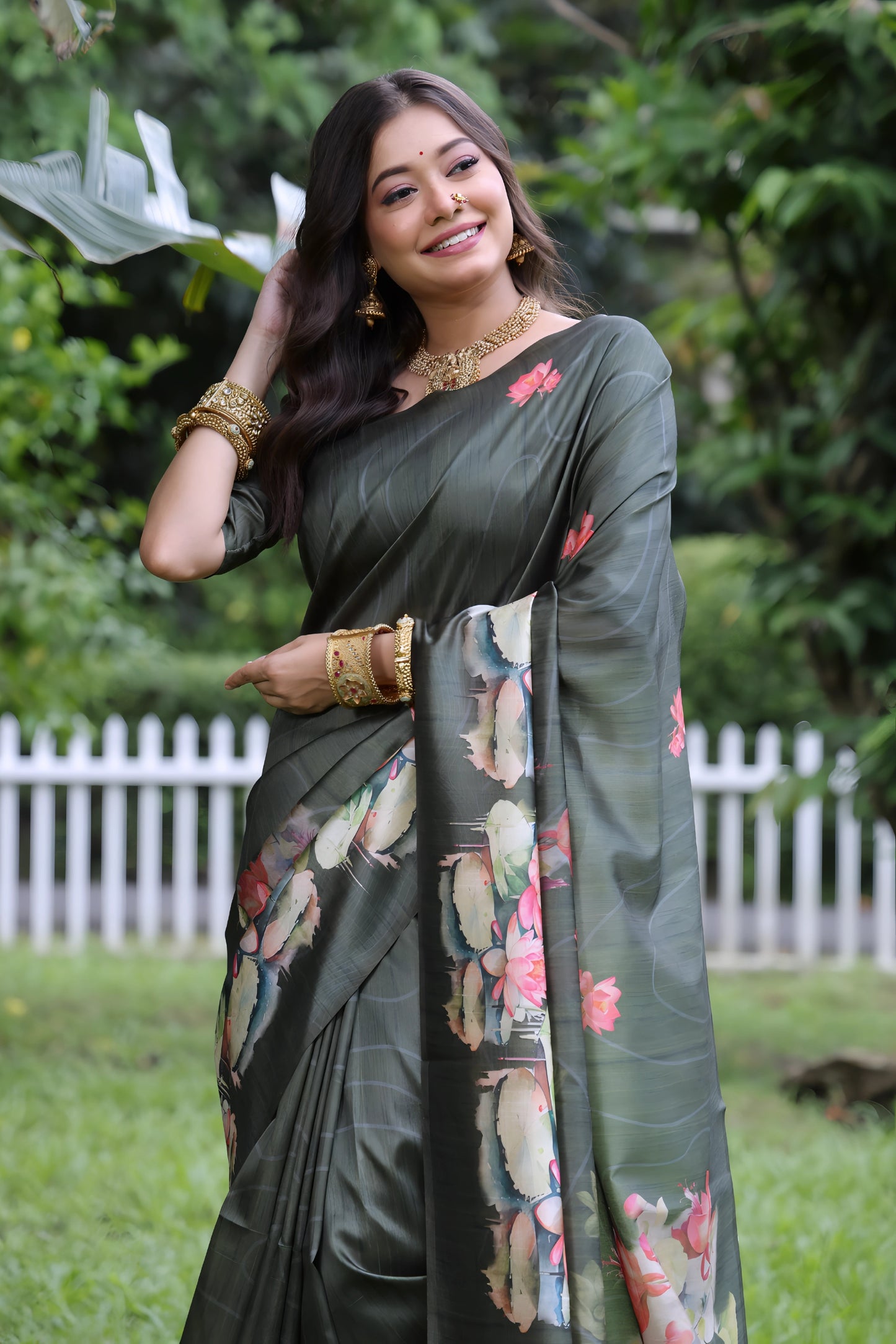 Woman wearing a dark green saree with large handpainted lotus and leaf floral motifs, accessorized with traditional gold jewelry and bangles, standing on a lush green lawn with tropical plants and a white picket fence in the background
