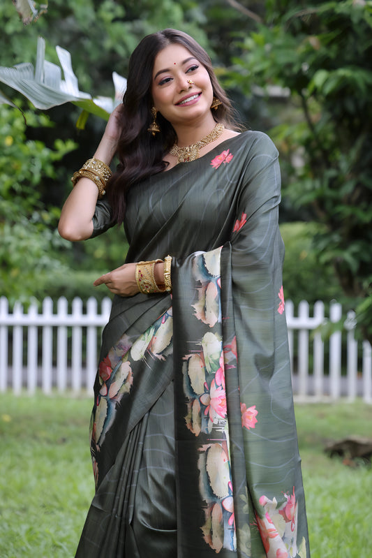 Woman wearing a dark green saree with large handpainted lotus and leaf floral motifs, accessorized with traditional gold jewelry and bangles, standing on a lush green lawn with tropical plants and a white picket fence in the background