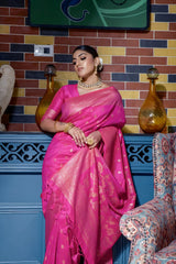 Woman wearing a hot pink silk saree with gold zari borders and polka dot motifs, paired with a matching blouse and traditional gold jewelry, elegantly posing in a vintage-inspired room with a blue cabinet, multicolored brick wall, artistic decor, and a patterned armchair.
