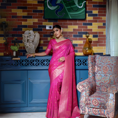 Woman wearing a hot pink silk saree with gold zari borders and polka dot motifs, paired with a matching blouse and traditional gold jewelry, elegantly posing in a vintage-inspired room with a blue cabinet, multicolored brick wall, artistic decor, and a patterned armchair.