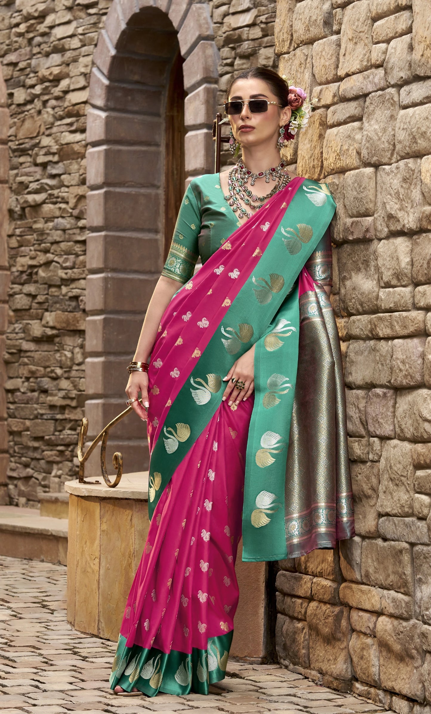 Woman in a pink and green saree standing against a stone wall.