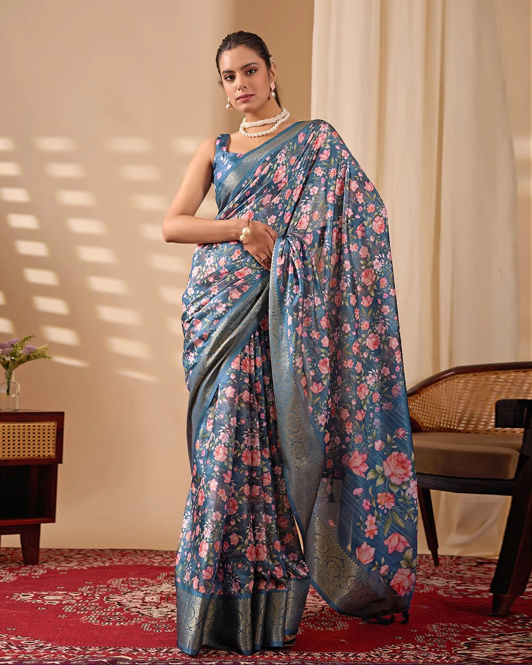 Woman wearing a blue floral saree in an indoor setting with a chair and table.