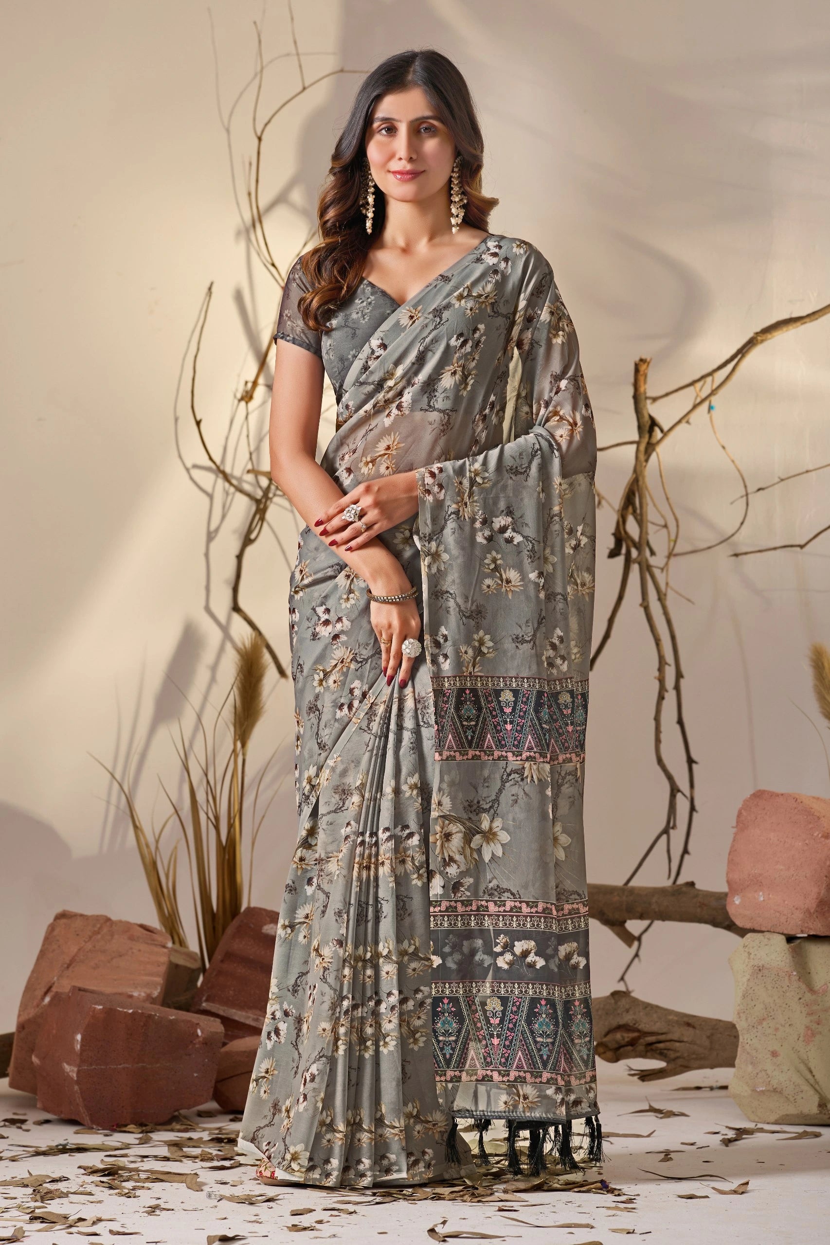 Woman wearing a patterned saree standing against a natural background with branches and rocks.