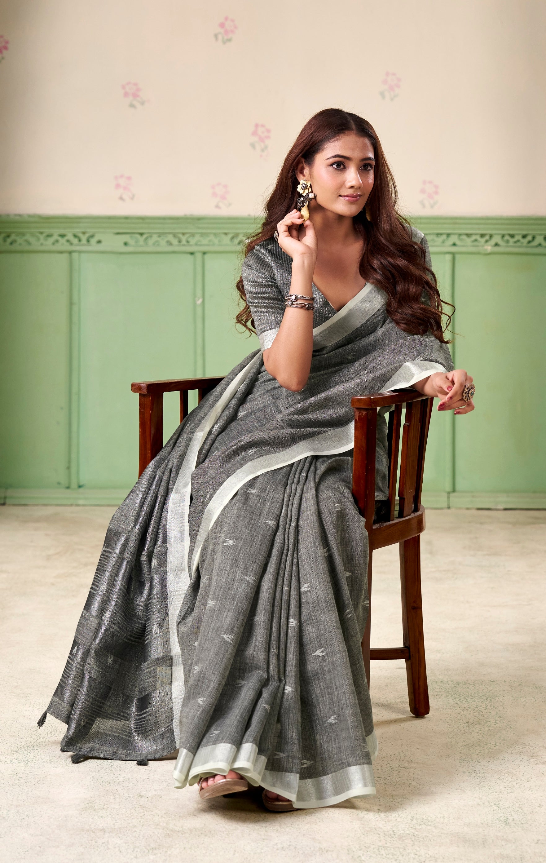 Woman in a gray saree sitting on a chair against a light green wall.