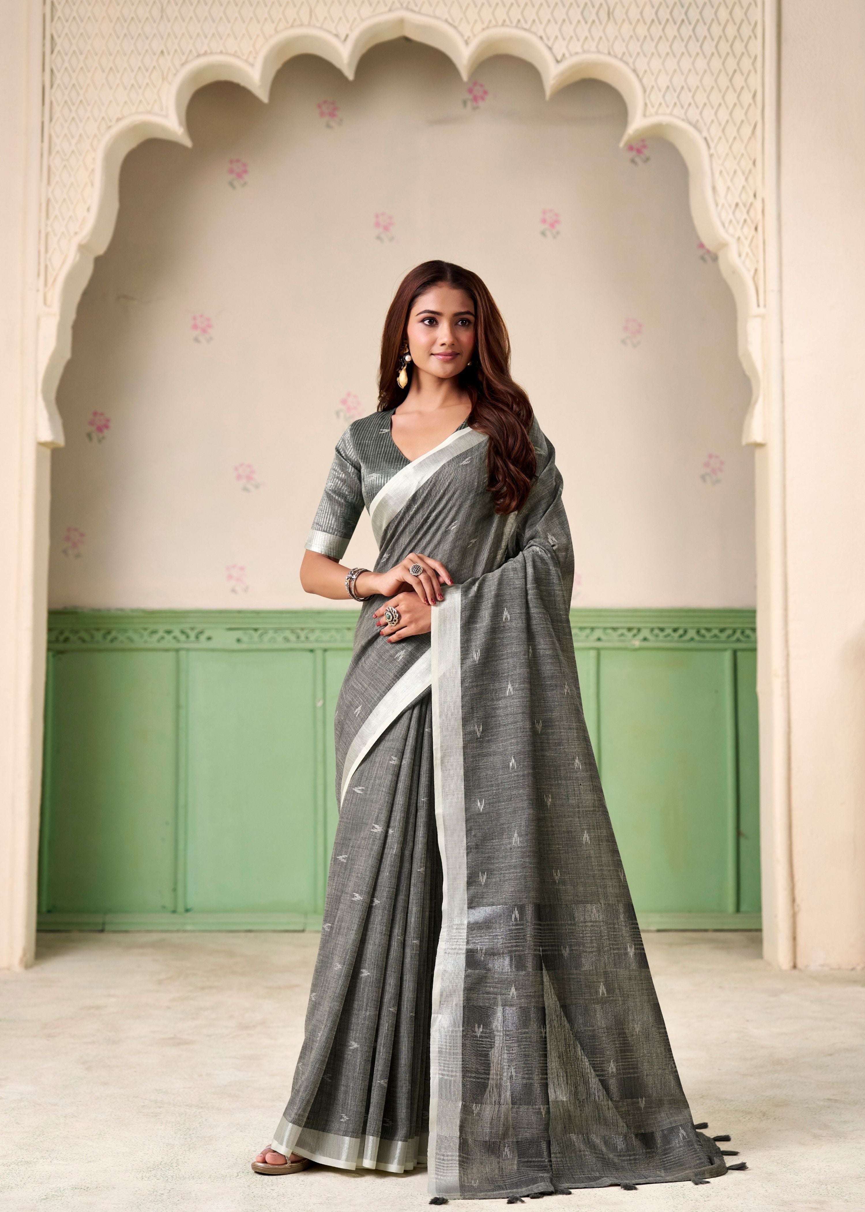 Woman wearing a gray saree with a white border indoors against a decorative wall.