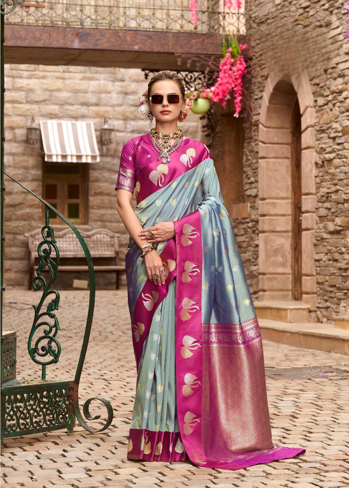Woman in a pink and blue saree standing in an outdoor setting with stone walls and decorative elements.