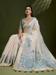 Woman wearing an ivory silk saree with grey fish and floral motifs, paired with a grey blouse and pearl jewelry, posing in a contemporary studio with olive green walls and a metal chair.