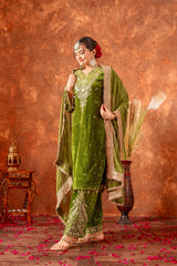 Woman in a green traditional outfit with gold embroidery against a textured brown background.