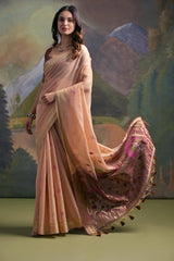 Woman wearing a beige silk saree with subtle floral embroidery and a maroon blouse, featuring a pallu adorned with purple floral motifs and tassels, standing in front of a painted mountain and forest backdrop, accessorized with traditional jewelry.