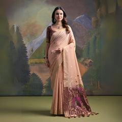 Woman wearing a beige silk saree with subtle floral embroidery and a maroon blouse, featuring a pallu adorned with purple floral motifs and tassels, standing in front of a painted mountain and forest backdrop, accessorized with traditional jewelry.