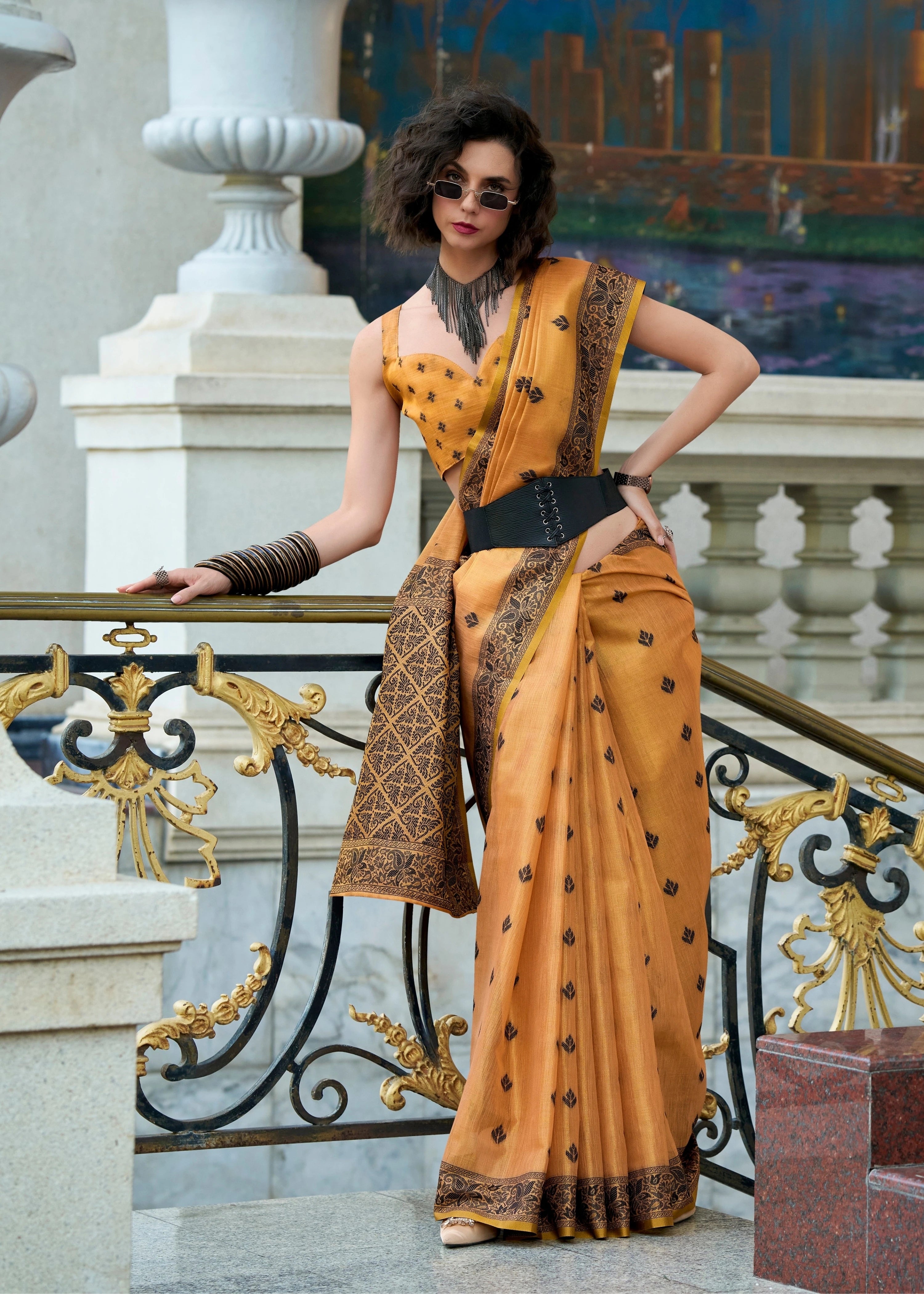 Woman in a mustard yellow saree with black patterns standing on a decorative staircase.