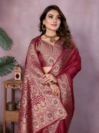 Woman wearing a mauve Banarasi silk saree adorned with an intricate golden zari border and detailed traditional motifs, paired with a matching blouse and gold jewelry. She stands gracefully in a modern studio featuring large rustic white clay pots, a wooden stand with brass decorative items, a bowl of colorful marigold flowers, and green foliage against a muted mauve backdrop.