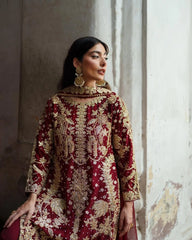 Woman wearing a red and gold embroidered traditional outfit against a textured wall.