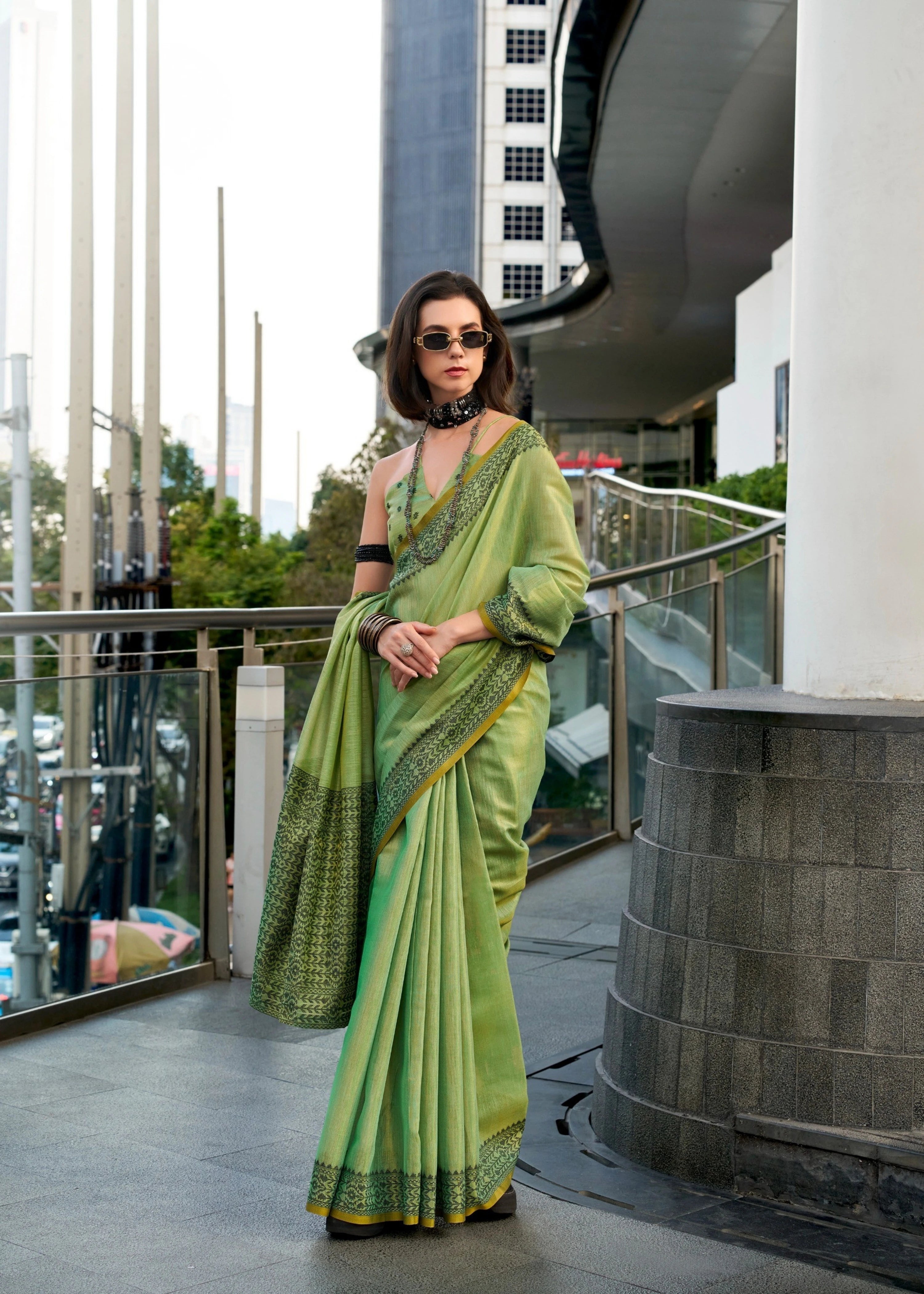 Woman in a green saree standing on a balcony with modern architecture in the background