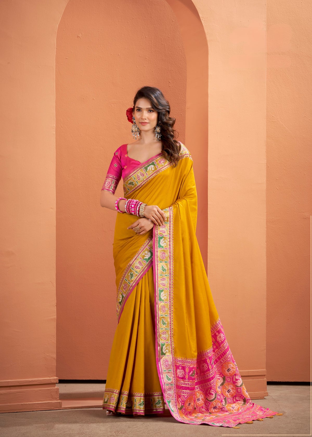 Woman wearing a yellow saree with pink blouse against a peach-colored wall.