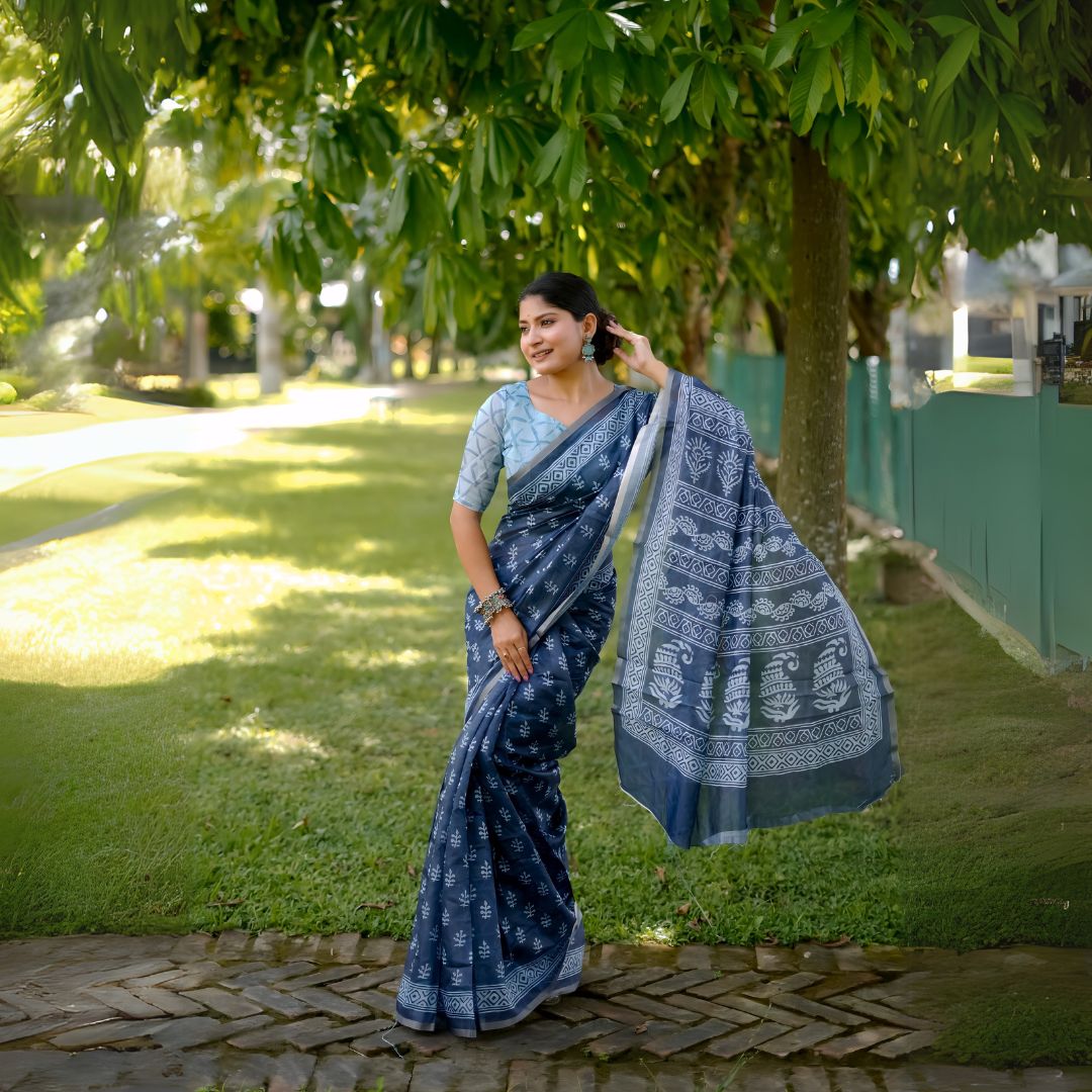 Woman wearing a blue handloom cotton saree with white tribal-inspired prints and a light blue blouse, accessorized with traditional jewelry, posing gracefully outdoors under lush green trees with sunlight filtering through the garden.