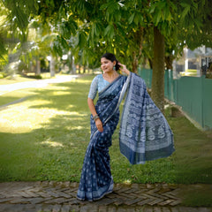 Woman wearing a blue handloom cotton saree with white tribal-inspired prints and a light blue blouse, accessorized with traditional jewelry, posing gracefully outdoors under lush green trees with sunlight filtering through the garden.