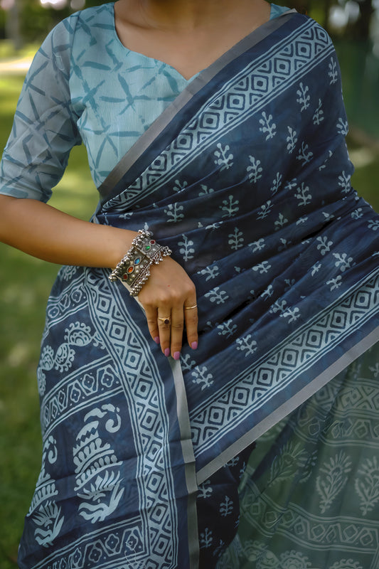 Woman wearing a blue handloom cotton saree with white tribal-inspired prints and a light blue blouse, accessorized with traditional jewelry, posing gracefully outdoors under lush green trees with sunlight filtering through the garden.