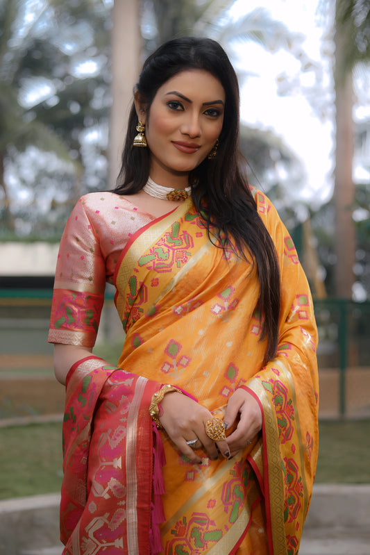 Woman wearing a yellow Banarasi silk saree adorned with gold zari motifs and a striking pink contrast pallu featuring traditional temple and floral patterns. She pairs the saree with a short sleeve pink blouse and accessorizes with gold jewelry, standing gracefully on a manicured lawn in an outdoor garden setting with fencing, lush greenery, and tropical trees in the background.