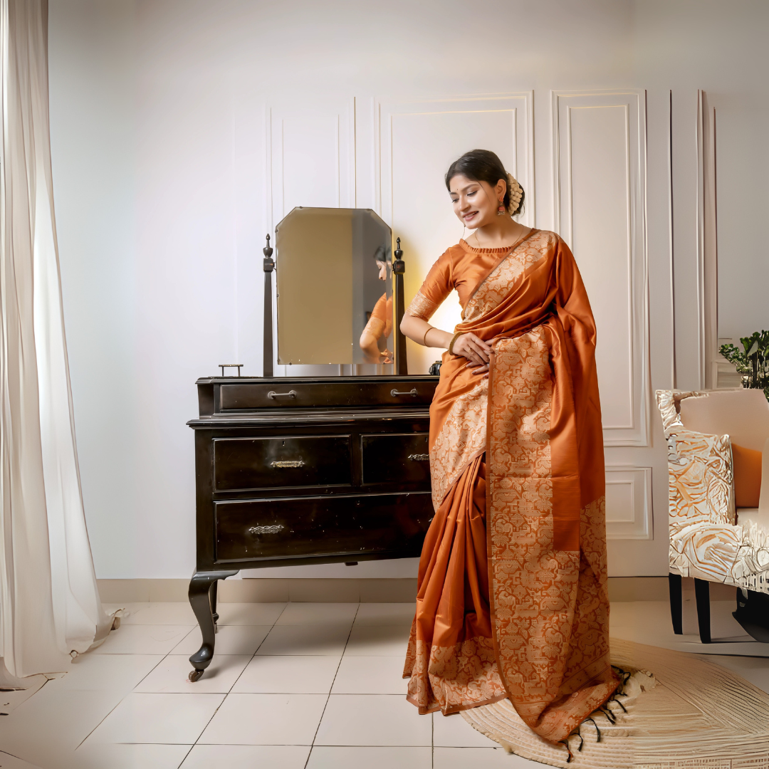 Woman wearing a copper silk saree with intricate brocade patterns and a matching blouse, standing elegantly beside a vintage black dresser with a mirror in a softly lit studio featuring white paneled walls and a patterned armchair.