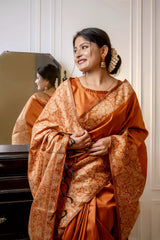 Woman wearing a copper silk saree with intricate brocade patterns and a matching blouse, standing elegantly beside a vintage black dresser with a mirror in a softly lit studio featuring white paneled walls and a patterned armchair.