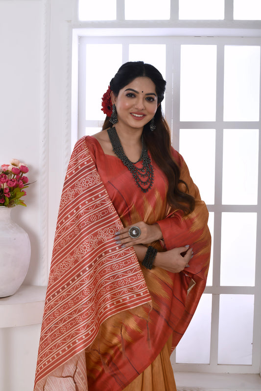 Woman wearing a beige and red Ikat printed saree with geometric patterns and a detailed pallu, paired with a red blouse and statement jewelry, standing in a modern white interior with floral decor and large arched window in the background.
