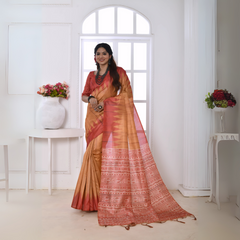 Woman wearing a beige and red Ikat printed saree with geometric patterns and a detailed pallu, paired with a red blouse and statement jewelry, standing in a modern white interior with floral decor and large arched window in the background.


