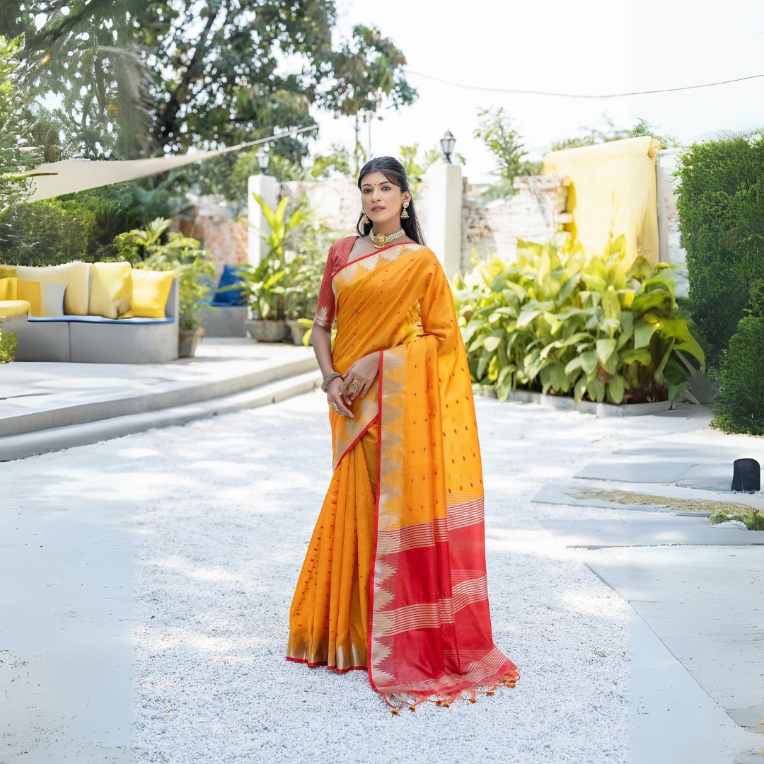 Woman wearing a vibrant yellow silk saree with a red and gold border, paired with a matching blouse and traditional jewelry, standing gracefully outdoors in a sunlit garden with lush green plants and modern seating in the background.