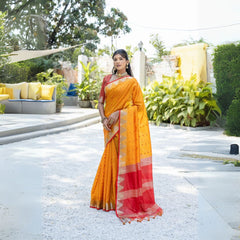 Woman wearing a vibrant yellow silk saree with a red and gold border, paired with a matching blouse and traditional jewelry, standing gracefully outdoors in a sunlit garden with lush green plants and modern seating in the background.