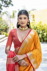 Woman wearing a vibrant yellow silk saree with a red and gold border, paired with a matching blouse and traditional jewelry, standing gracefully outdoors in a sunlit garden with lush green plants and modern seating in the background.