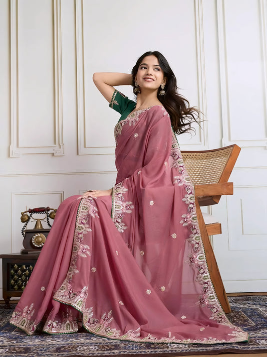  Woman wearing a dusty rose designer saree adorned with sequin detailing and an ornate embroidered floral border, paired with a contrasting green blouse embellished with small floral motifs. She stands elegantly in a classic white paneled room featuring a wooden armchair with a woven seat and a vintage rotary telephone on a carved wooden side table, all set atop an intricate patterned rug for a refined, traditional look.